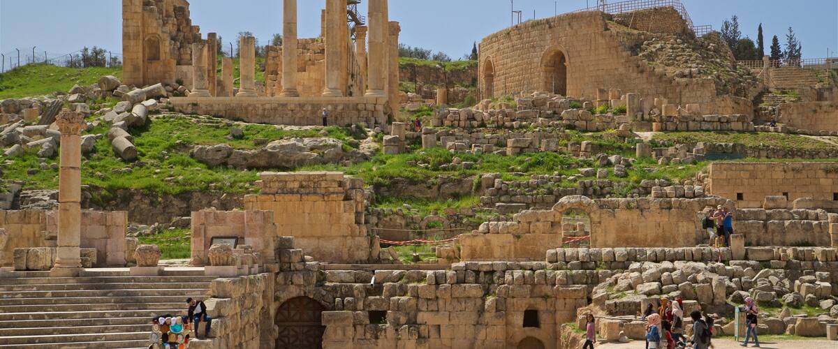 Ancient ruins with columns and arches, a staircase, and a hillside with greenery.