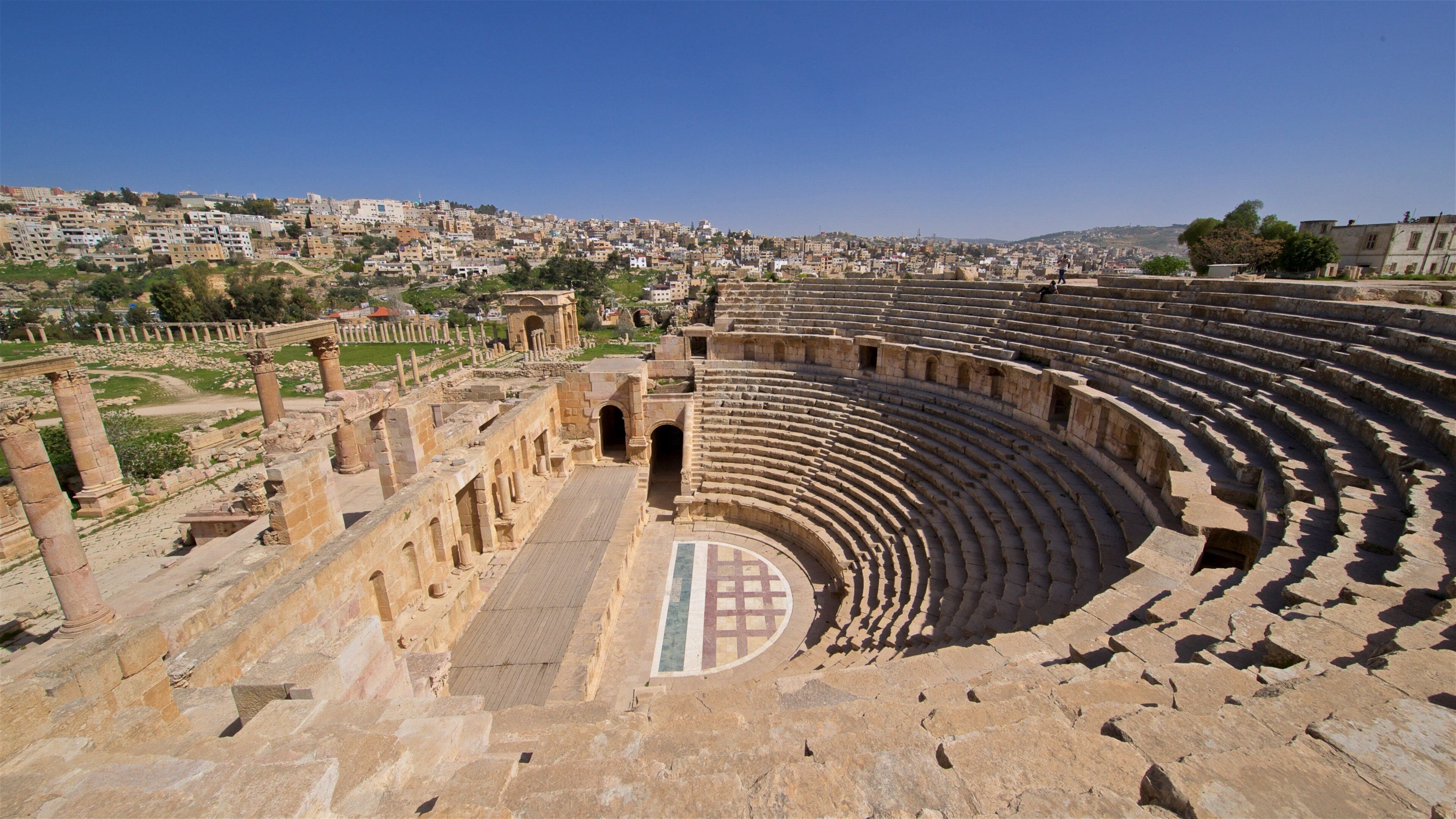 An ancient amphitheater with stone seating and columns, surrounded by ruins and a cityscape in the background.