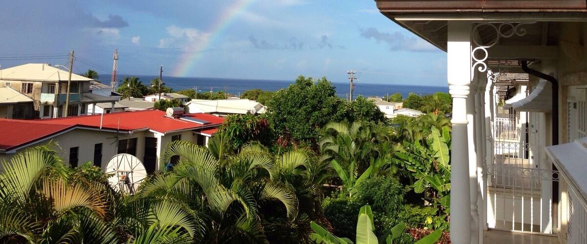 A house with a red roof, a white porch, and a view of the ocean with a rainbow.