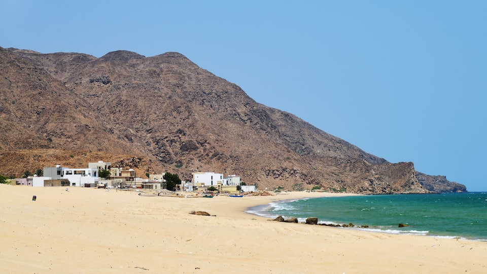 A sandy beach with buildings and a rocky mountain in the background.