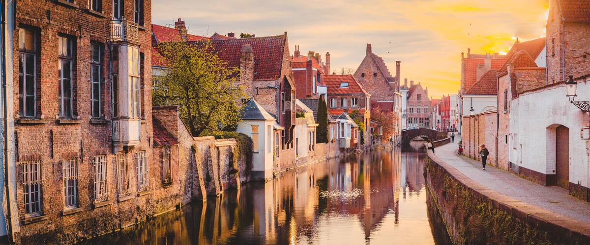 A canal with brick buildings on either side, a bridge in the distance, and a sunset sky.