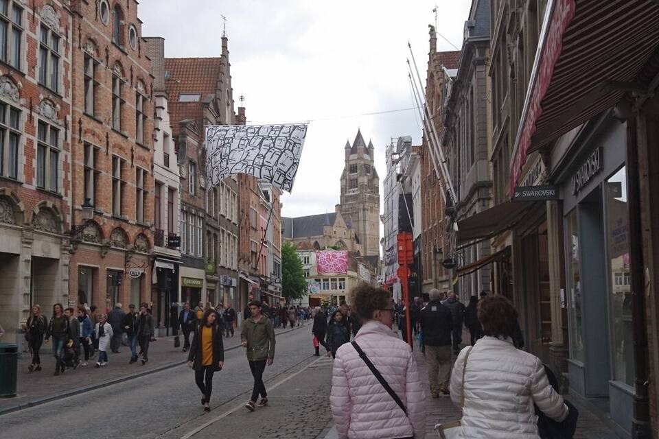 A cobblestone street lined with historic buildings, a prominent tower in the background, and pedestrians walking along the sidewalk.