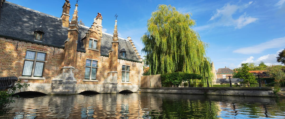 A historic brick building with multiple chimneys and a weeping willow tree beside a canal.