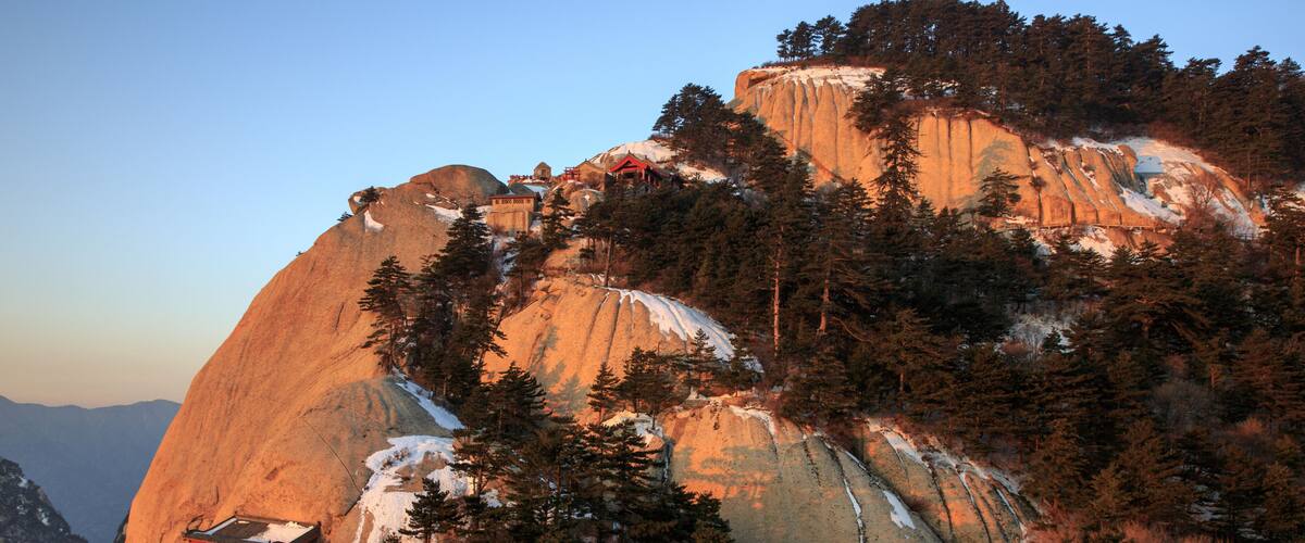 A mountain with a large rock formation, a small building, and dense forest.