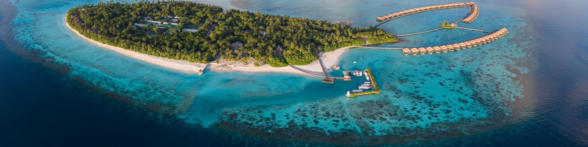 Vista aérea de una isla tropical con una playa, un resort con bungalows sobre el agua y aguas turquesas cristalinas.