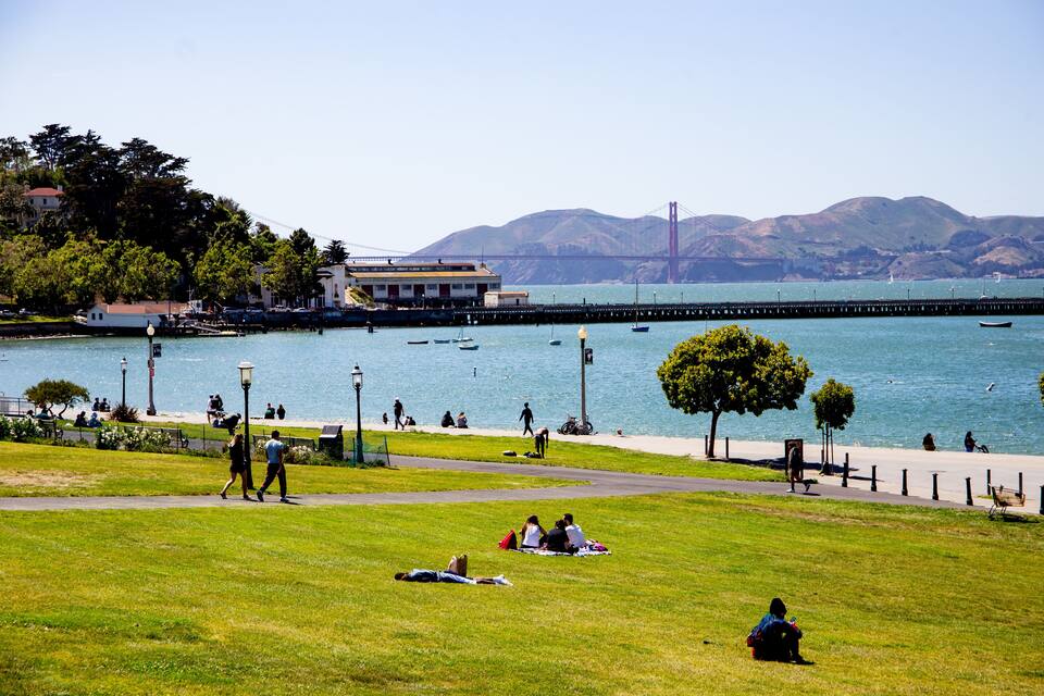 A park with people walking and sitting on the grass, a body of water, boats, and a bridge in the background.