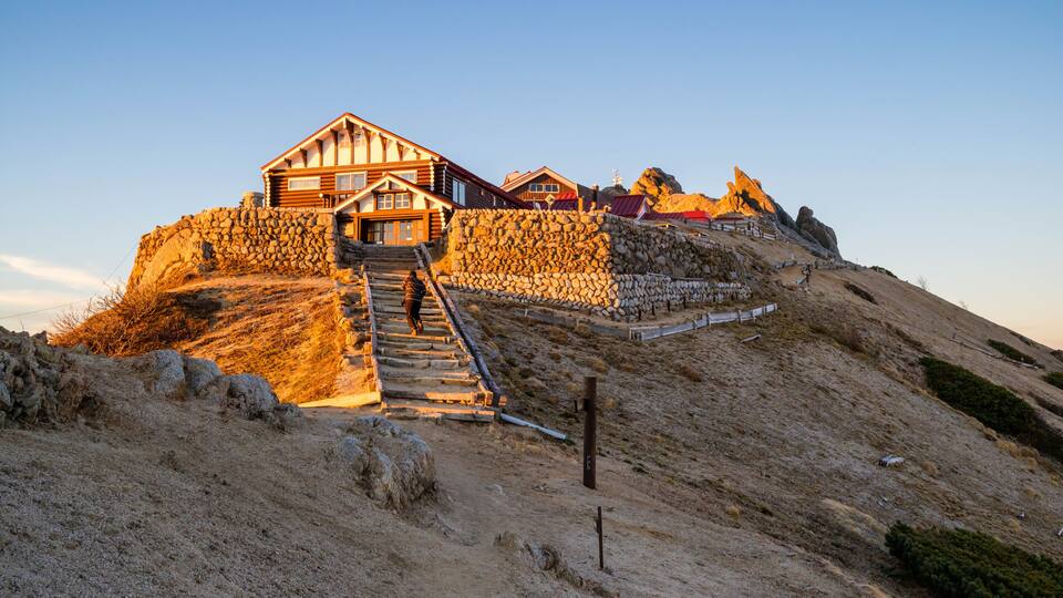 A wooden cabin with a stone staircase leading up to it, surrounded by a rocky hillside.