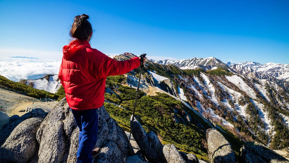 A person in a red jacket standing on rocky terrain with a view of snow-capped mountains and lush greenery.