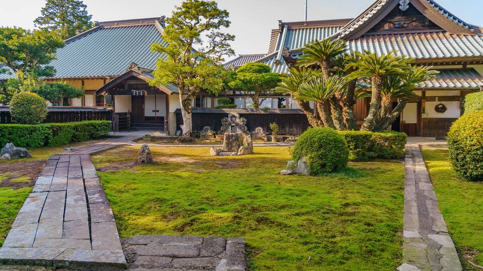A traditional Japanese garden with stone lanterns, a wooden walkway, and a building with a tiled roof.