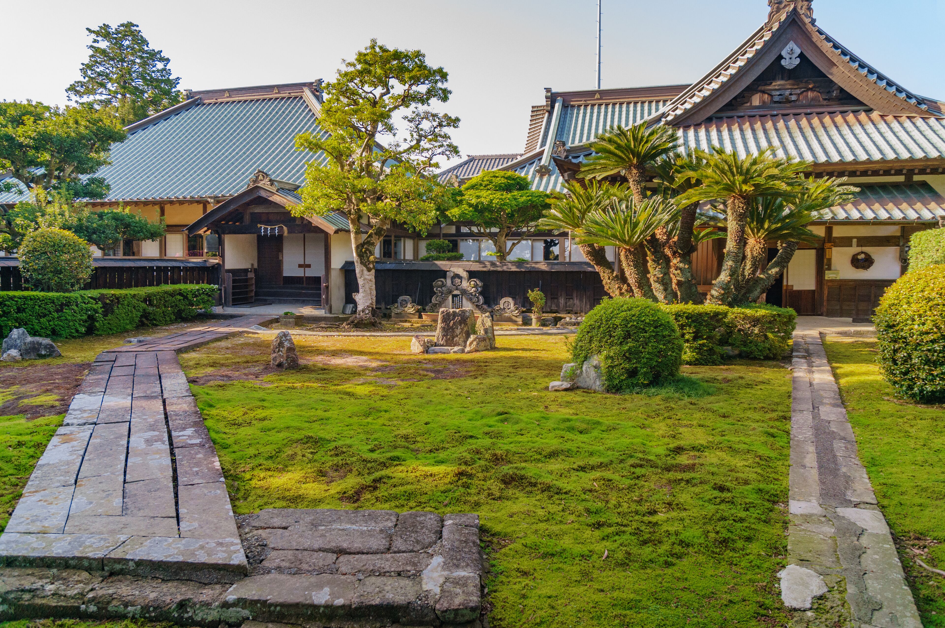 A traditional Japanese garden with stone lanterns, a wooden walkway, and a building with a tiled roof.