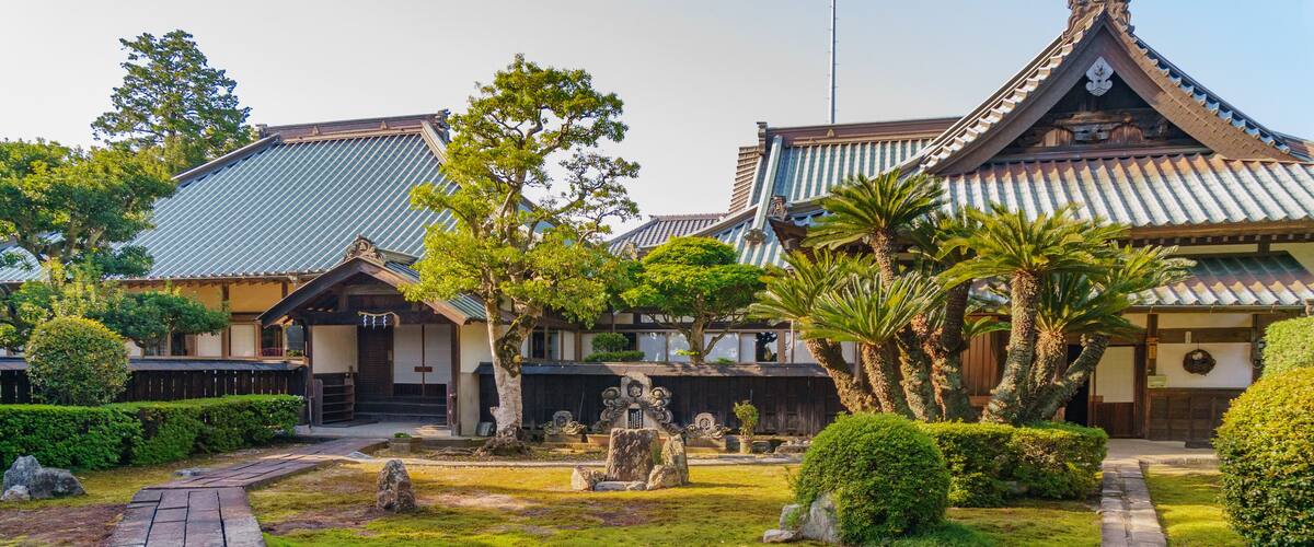A traditional Japanese garden with stone lanterns, a wooden walkway, and a building with a tiled roof.