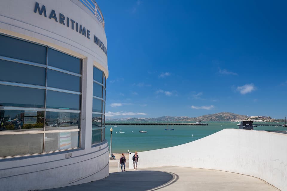 A modern building with a large glass facade and the words "MARITIME MUSEUM" on it. In the background, there is a clear blue sky, calm sea, and a distant hilly landscape.