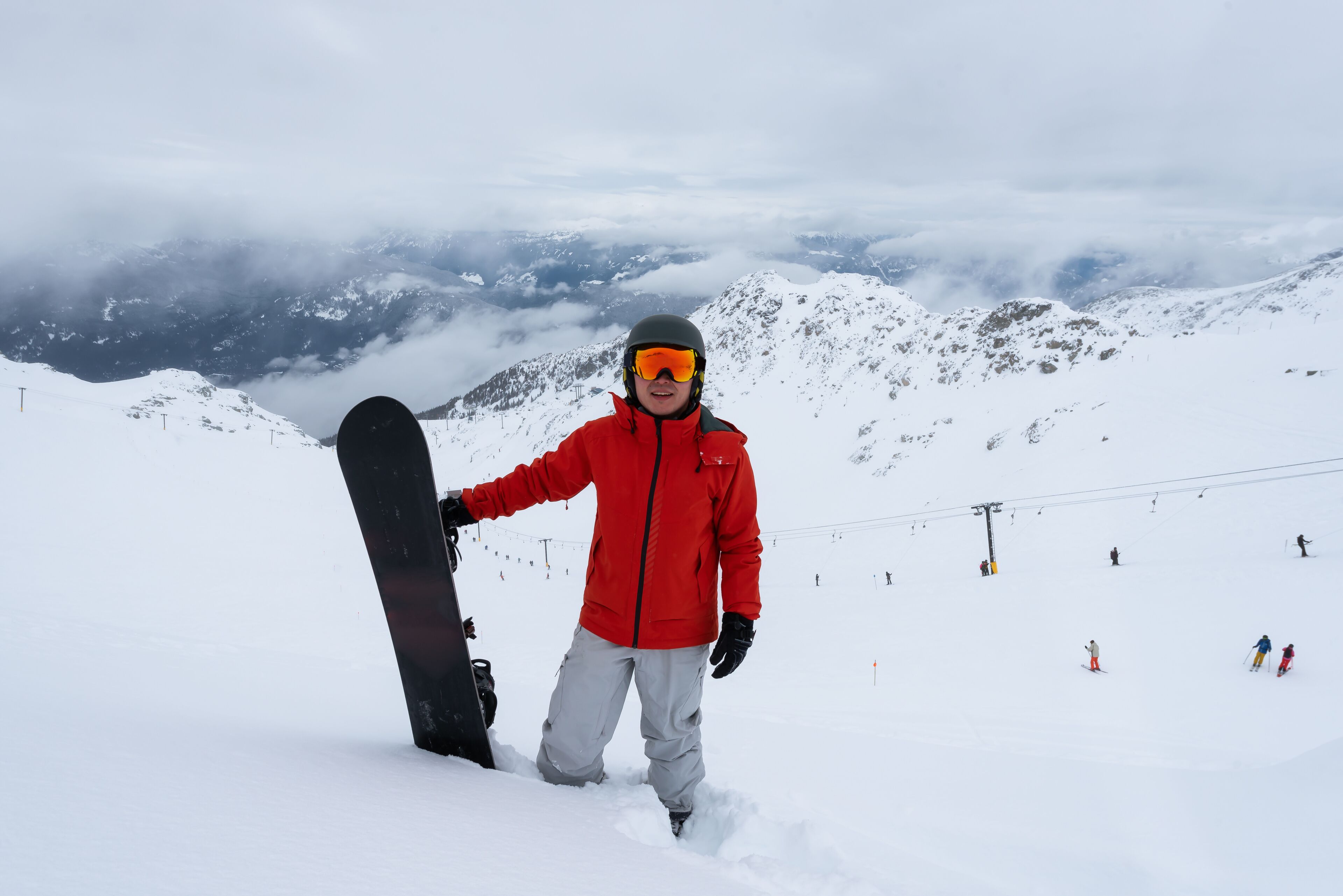 A person in a red jacket holding a snowboard on a snowy mountain with ski slopes in the background.
