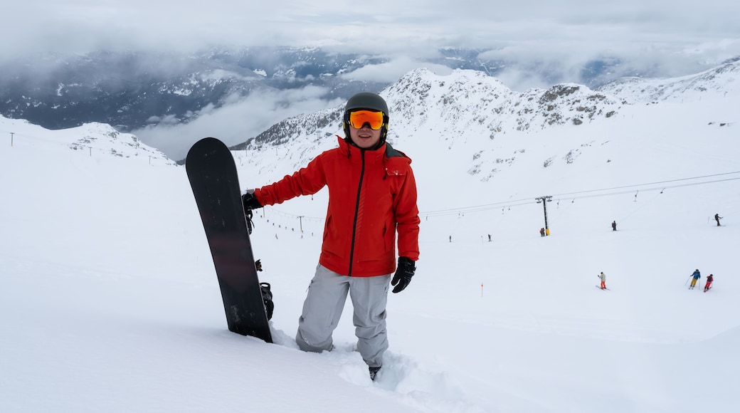A person in a red jacket holding a snowboard on a snowy mountain with ski slopes in the background.