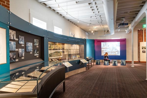 An indoor exhibition space with glass display cases, framed photographs, and a projection screen.