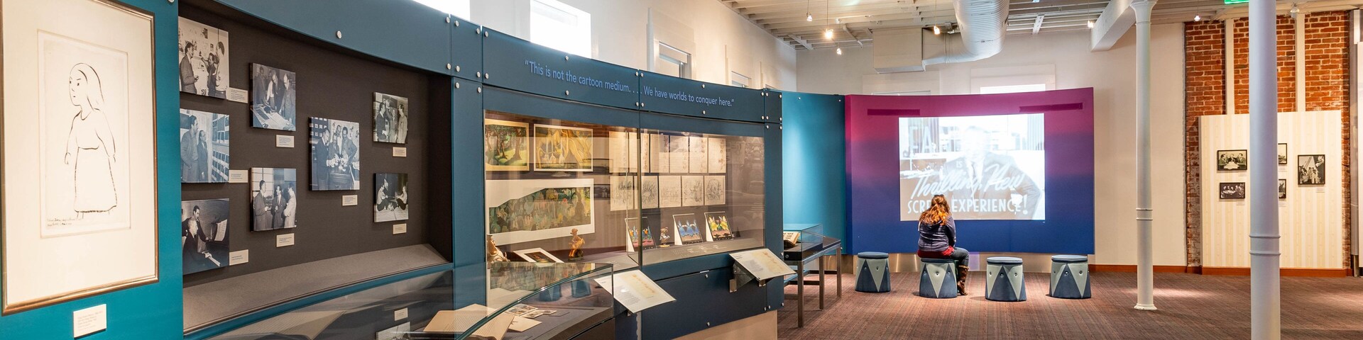 An indoor exhibition space with glass display cases, framed photographs, and a projection screen.