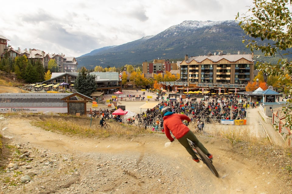 A mountain biking event with spectators, a dirt path, and buildings in the background.
