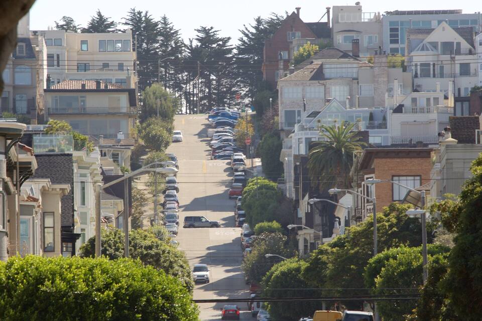 Rua íngreme, bordeada por árvores, com carros estacionados e edifícios residenciais.