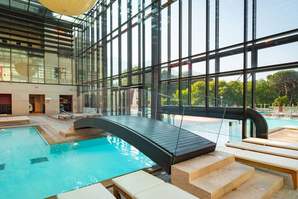 An indoor swimming pool with clear glass walls, wooden benches, and a glass bridge.