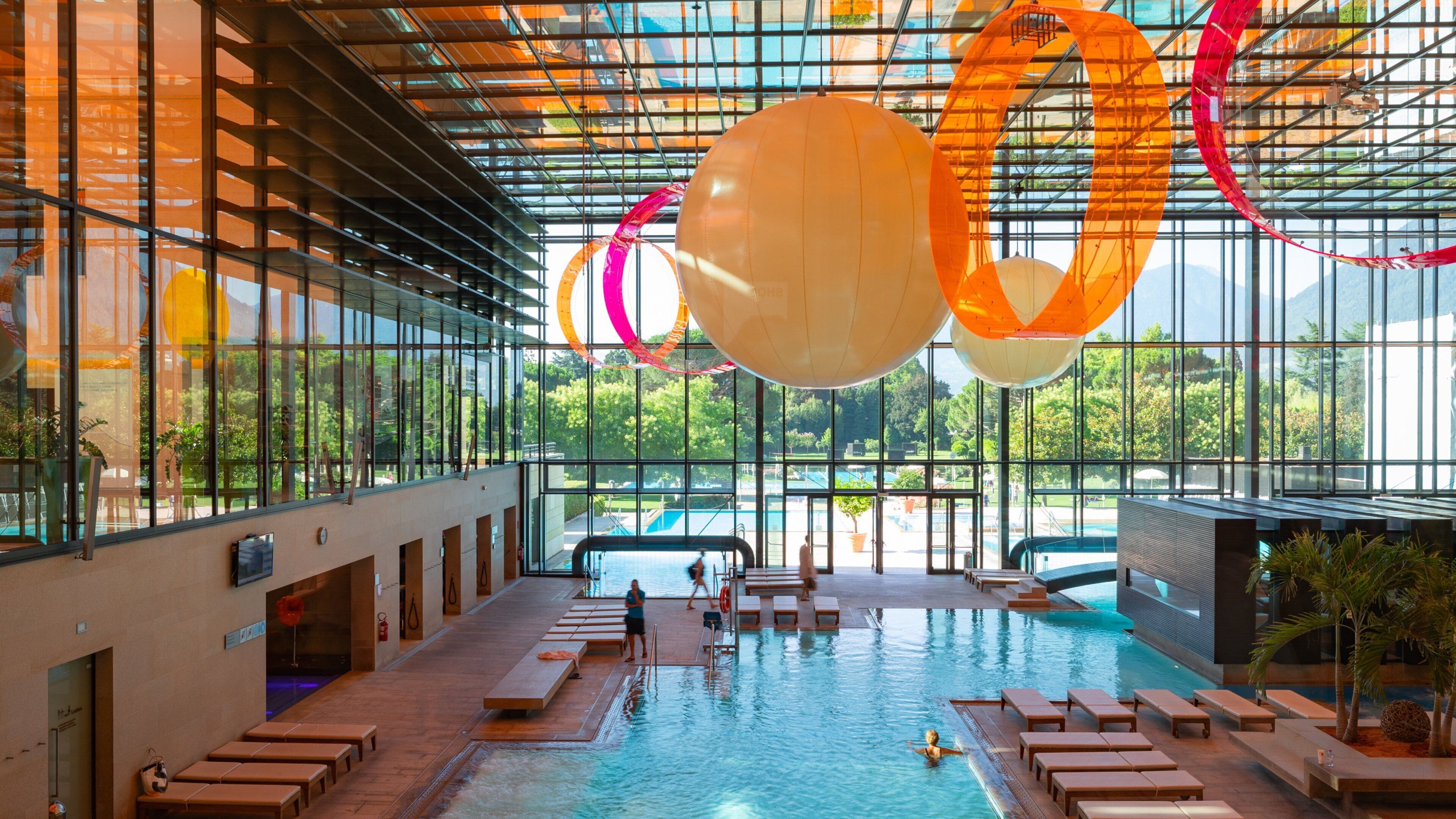 Une piscine intérieure dotée de grandes fenêtres, de chaises longues et d’éléments décoratifs suspendus.