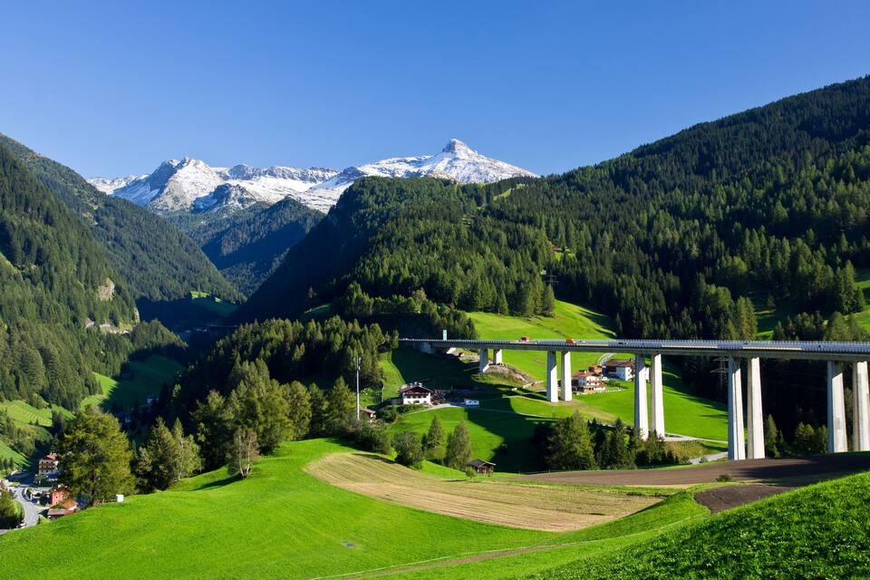 A mountainous landscape with a bridge, green fields, and a snow-capped peak.