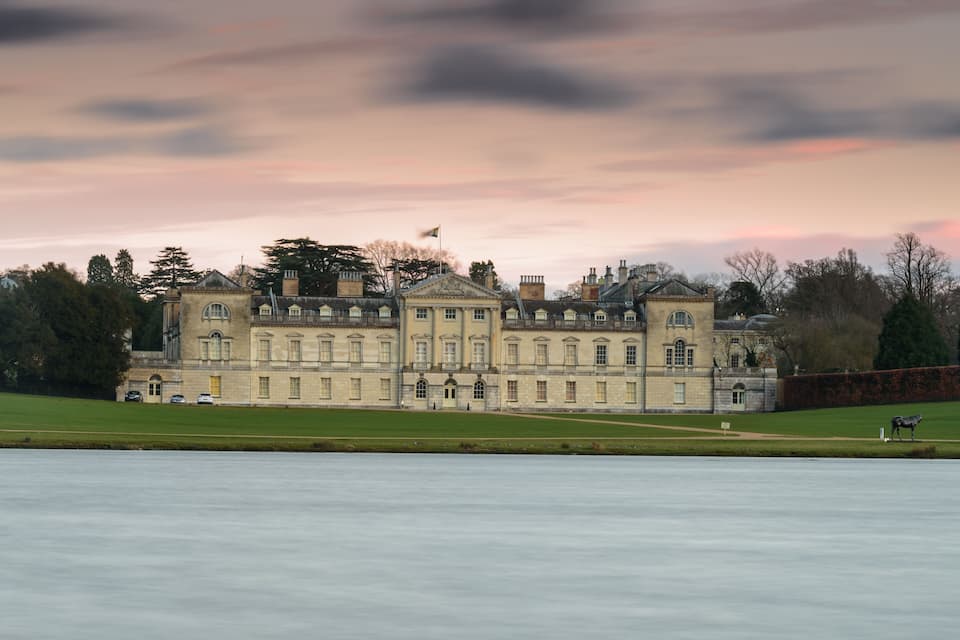 Un grand bâtiment imposant, doté d’une architecture classique, avec une pelouse parfaitement entretenue et un plan d’eau paisible en premier plan.