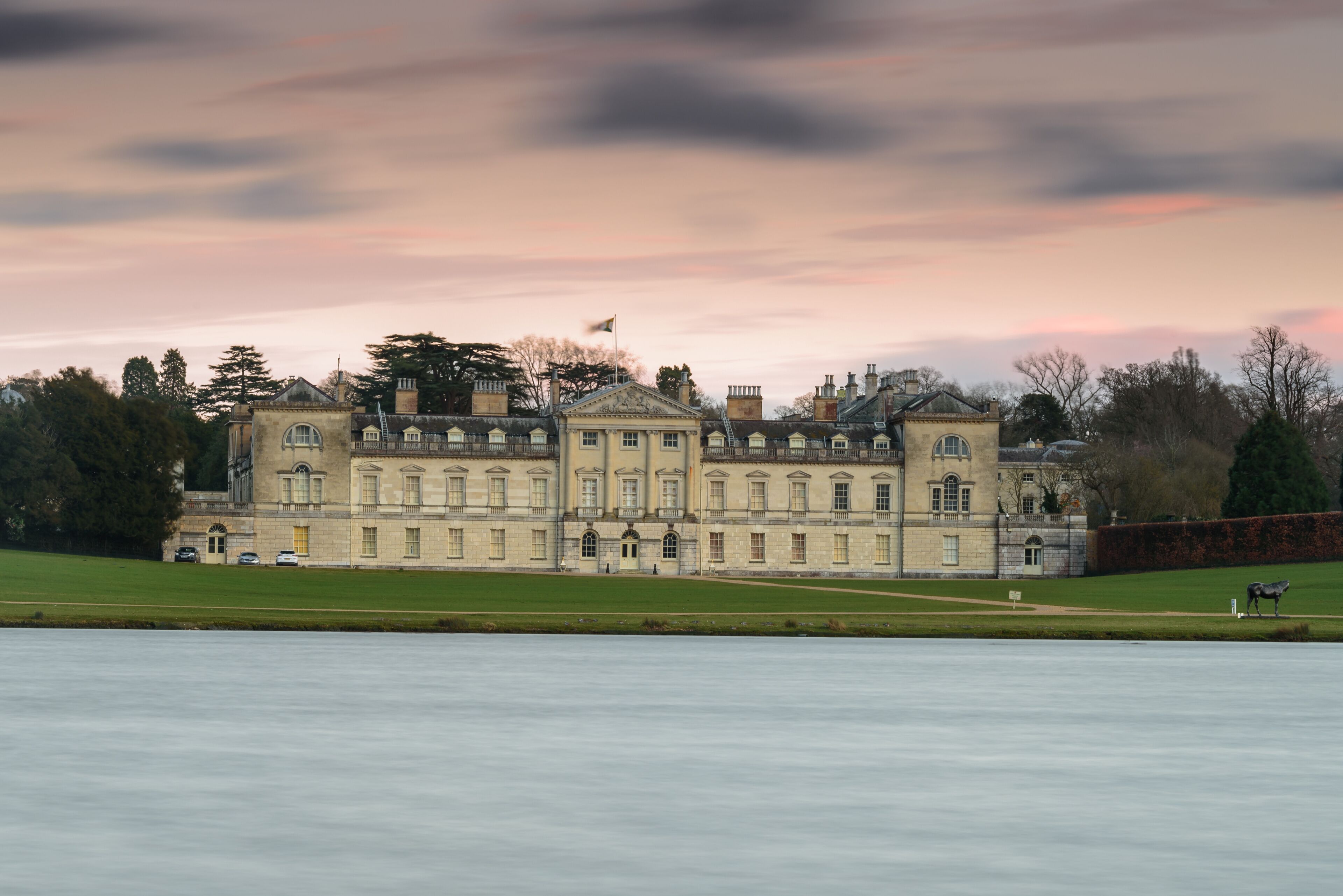 A large stately building with classical architecture, a manicured lawn, and a calm body of water in the foreground.