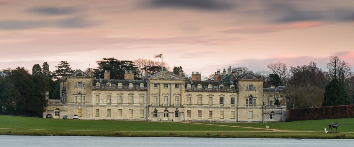 A large stately building with classical architecture, a manicured lawn, and a calm body of water in the foreground.