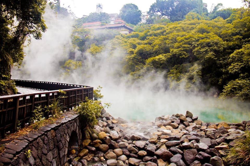 A stone bridge over a rocky area with a misty hot spring and lush greenery in the background.