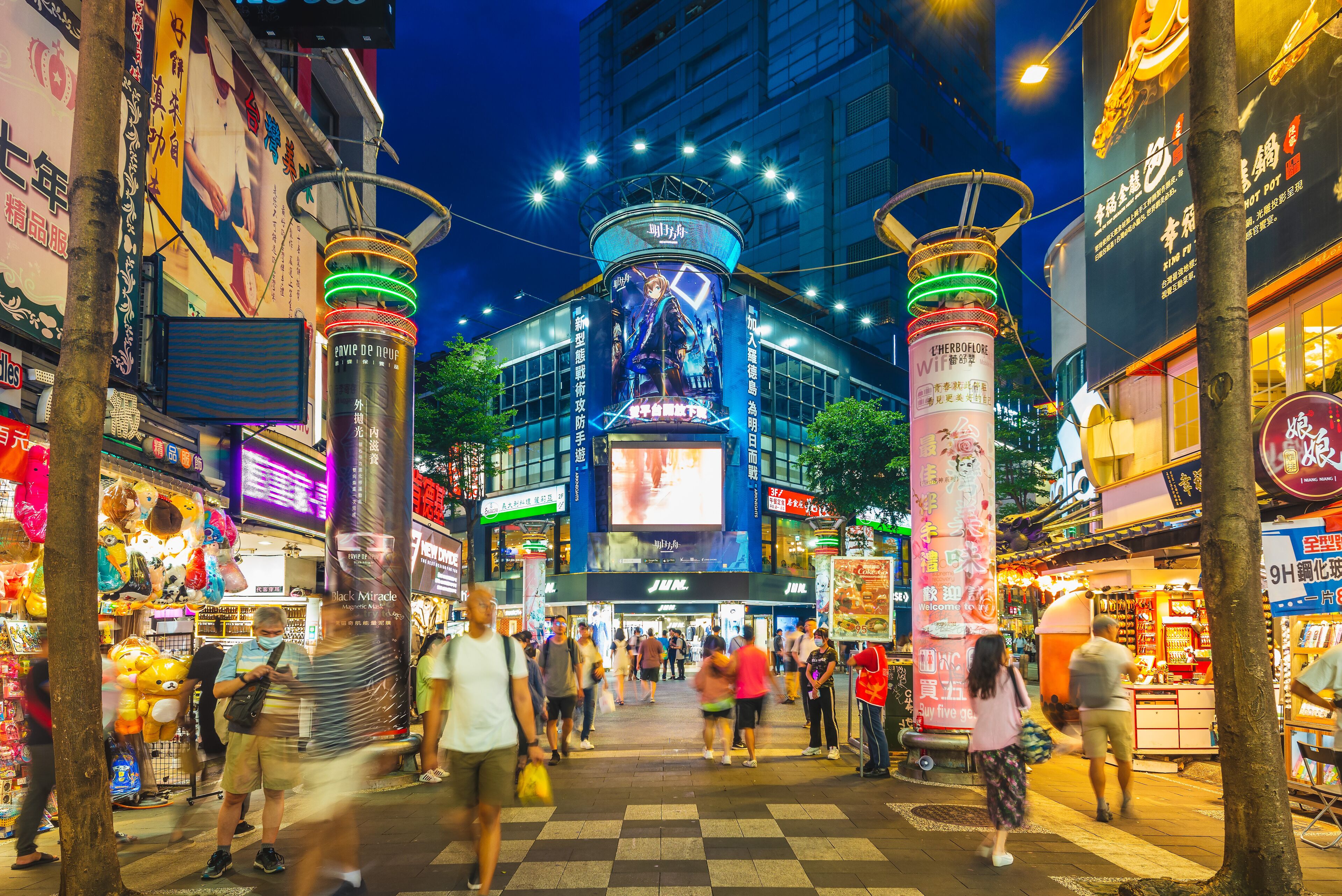 A bustling urban street at night with illuminated signage, a prominent digital billboard, and a variety of shops and eateries.