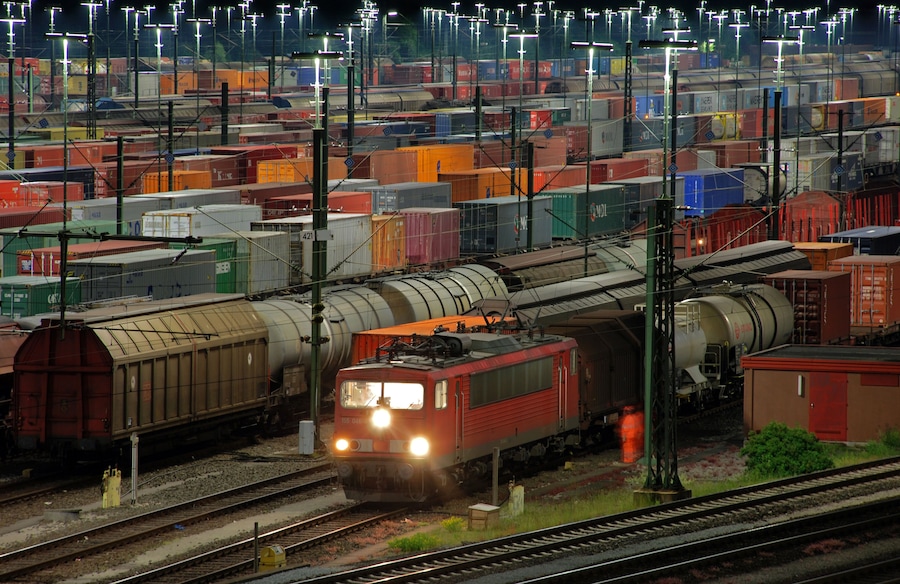 Ein Güterzug durchquert einen belebten Rangierbahnhof mit zahlreichen Gleisen und vielen Containern.
