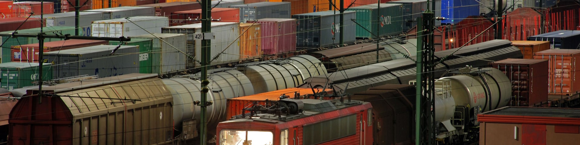 Ein Güterzug durchquert einen belebten Rangierbahnhof mit zahlreichen Gleisen und vielen Containern.