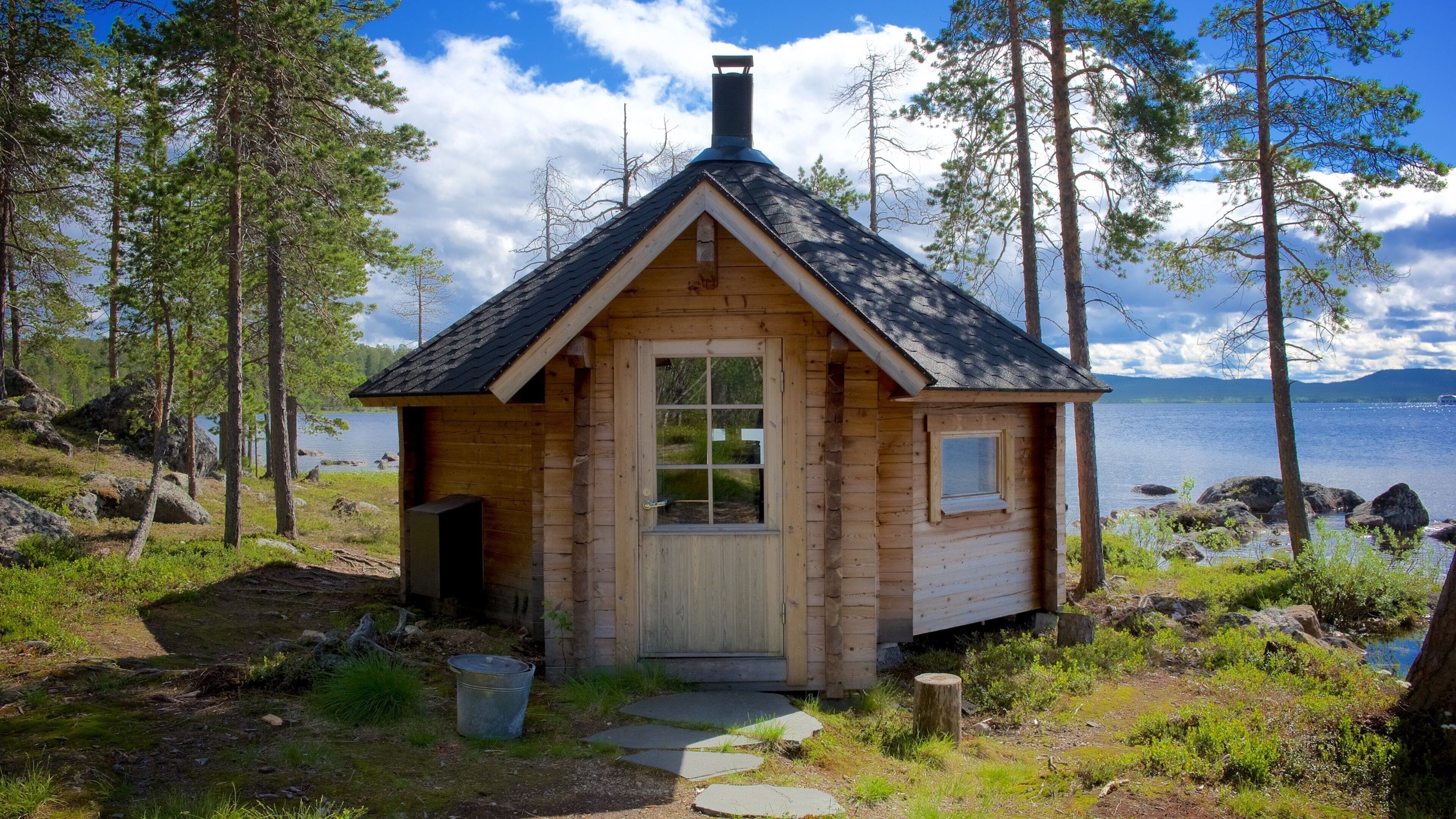 Pieni puinen mökki, jossa on kalteva katto, ovi ja ikkuna, ja sen ympärillä on puita ja järvi.