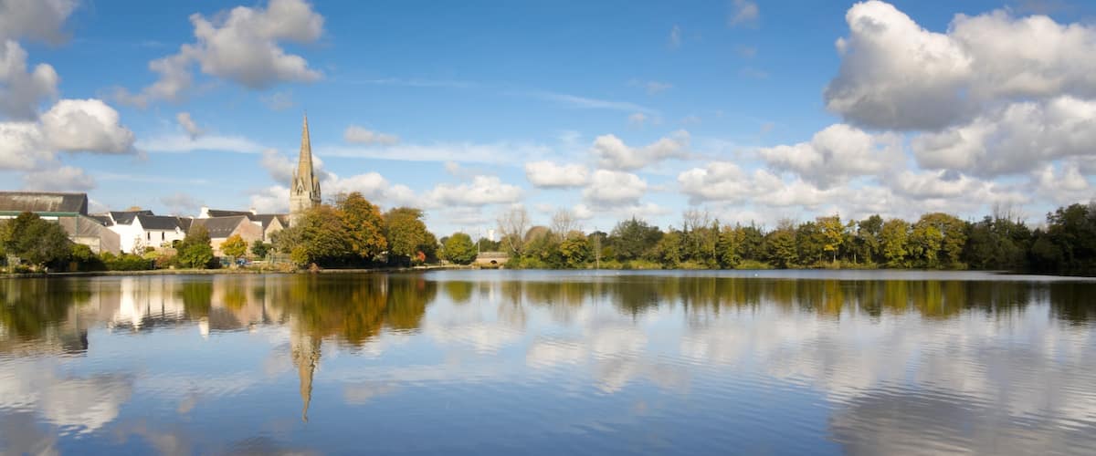A calm lake with a church spire, trees, and buildings reflecting in the water.