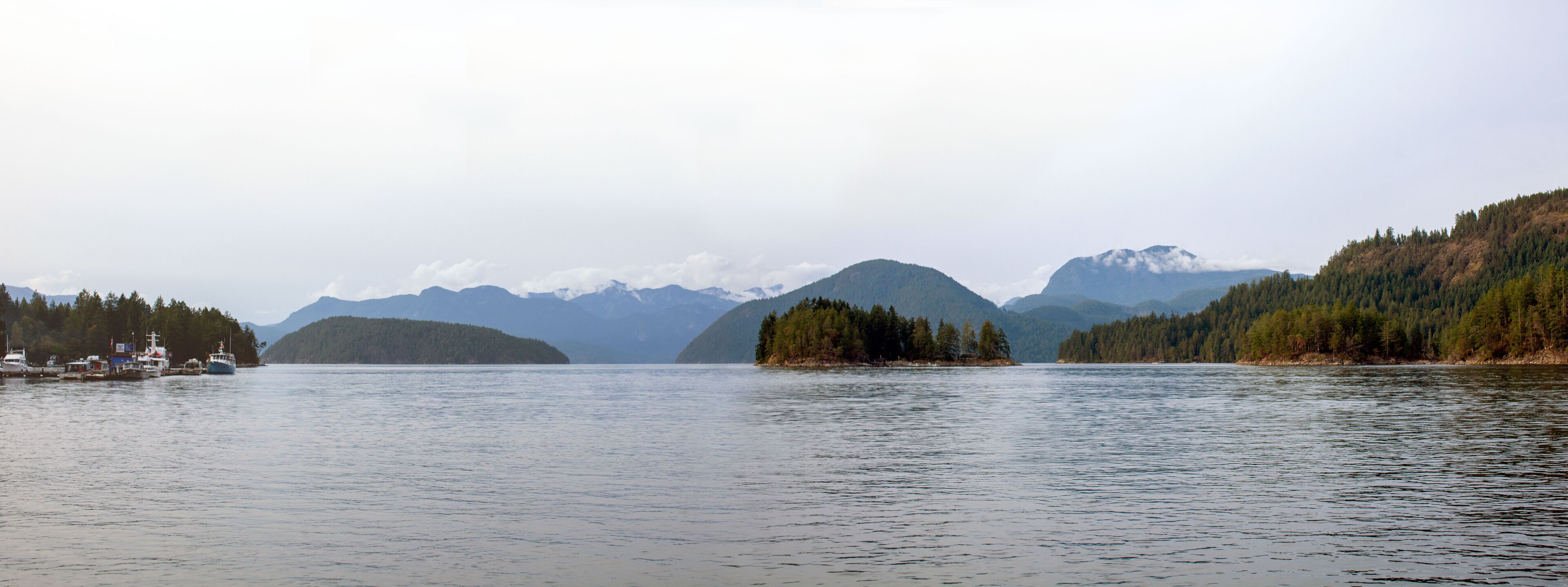 A calm lake with boats, surrounded by forested hills and distant mountains.