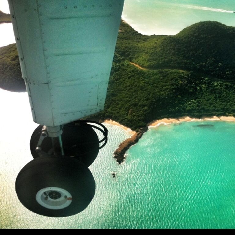 An aerial view of a coastal area with turquoise waters, a sandy beach, and lush green hills.