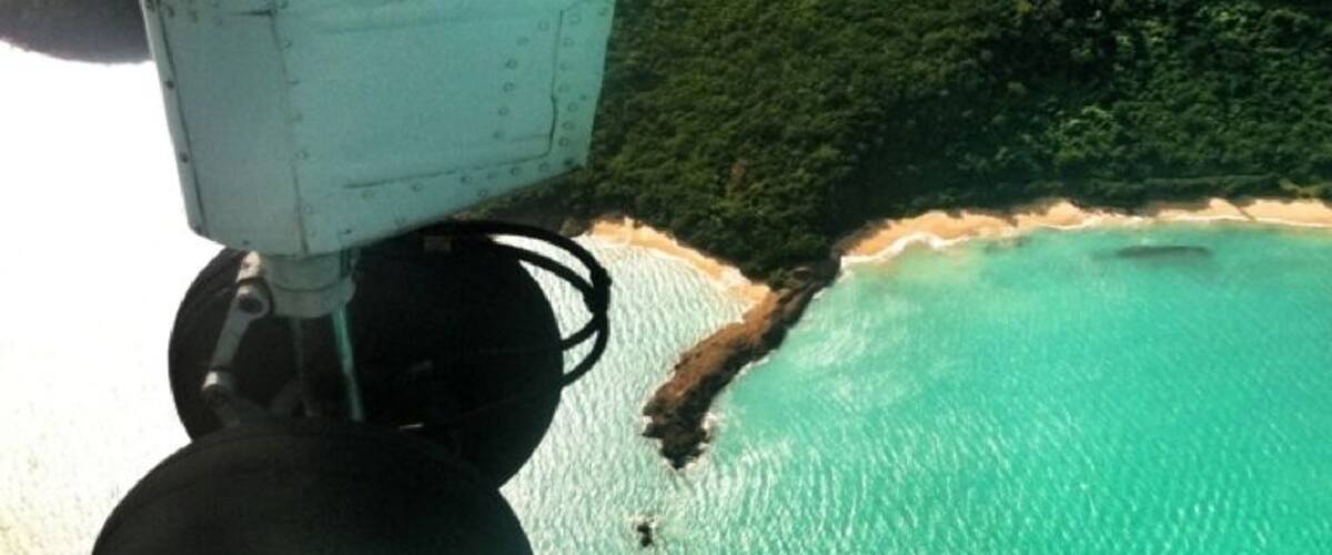 An aerial view of a coastal area with turquoise waters, a sandy beach, and lush green hills.
