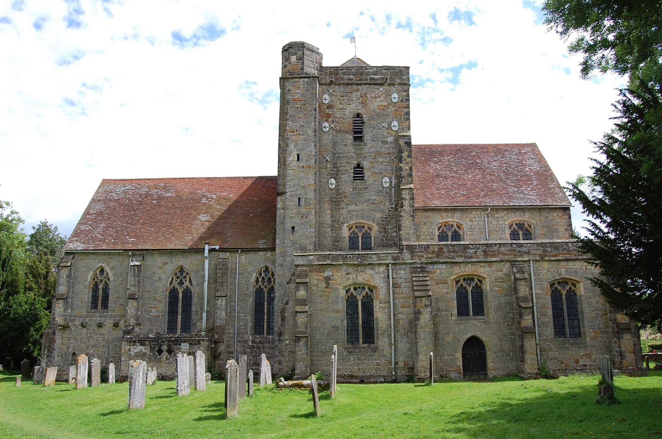 Late 14th Century church with axial tower, north and south aisles and short nave, very little altered from the original church built by Sir William De Etchyngham who died in 1389. His memorial brass is in pride of place in front of the altar. The tower has a rare 14th C. weather vane on top, depicting the arms of the De Etchingham family. There is one bell. 14th C. south porch and 13th C. font, which may pre-date the church, as a chapel stood on the site previously. Originally the De Etchingham's manor stood nearby (probably under the railway station) and traces of the moat can still be discerned in the churchyard. Lower part of the rood screen is original, as are the medieval encaustic floor tiles. There was once a chantry chapel or sacristy on the north side of the chancel, which is now gone, although a piscina and blocked door can still be seen. The choir stalls date from about 1375, and retain the original misericords, with matching designs on north and south sides. A series of vigorous carvings show dolphins, keys, lady's heads and a fox preaching to geese. The stalls are raised slightly on a stone platform. There is a fine triple sedilia on the south side of the chancel with the usual piscina. The chancel floor has several wonderful brasses - Sir William shown in armour with feet on a lion. His head and the escutcheons are missing. West of this is a fine triple canopied brass to Sir William's son - also William, his wife Joan and son Thomas, who died in 1444. The brasses were made in London. The church was carefully restored by William Slater around 1856, but by the early 20th C. the tower was in a perilous state, the inherent design of a central tower was always difficult, and subsidence had occurred. It was found there were virtually no foundations! In 1938 tower underpinning and tie-bars were used to stabilise the tower and walls. In 2000 the church was badly flooded as it stands near the Rother and Dudwell rivers. Water reached up to the base of the communion rail. Luckily insurance paid for much of the clean up and restoration.