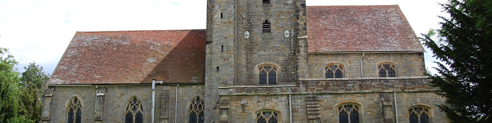 Late 14th Century church with axial tower, north and south aisles and short nave, very little altered from the original church built by Sir William De Etchyngham who died in 1389. His memorial brass is in pride of place in front of the altar. The tower has a rare 14th C. weather vane on top, depicting the arms of the De Etchingham family. There is one bell. 14th C. south porch and 13th C. font, which may pre-date the church, as a chapel stood on the site previously. Originally the De Etchingham's manor stood nearby (probably under the railway station) and traces of the moat can still be discerned in the churchyard. Lower part of the rood screen is original, as are the medieval encaustic floor tiles. There was once a chantry chapel or sacristy on the north side of the chancel, which is now gone, although a piscina and blocked door can still be seen. The choir stalls date from about 1375, and retain the original misericords, with matching designs on north and south sides. A series of vigorous carvings show dolphins, keys, lady's heads and a fox preaching to geese. The stalls are raised slightly on a stone platform. There is a fine triple sedilia on the south side of the chancel with the usual piscina. The chancel floor has several wonderful brasses - Sir William shown in armour with feet on a lion. His head and the escutcheons are missing. West of this is a fine triple canopied brass to Sir William's son - also William, his wife Joan and son Thomas, who died in 1444. The brasses were made in London. The church was carefully restored by William Slater around 1856, but by the early 20th C. the tower was in a perilous state, the inherent design of a central tower was always difficult, and subsidence had occurred. It was found there were virtually no foundations! In 1938 tower underpinning and tie-bars were used to stabilise the tower and walls. In 2000 the church was badly flooded as it stands near the Rother and Dudwell rivers. Water reached up to the base of the communion rail. Luckily insurance paid for much of the clean up and restoration.