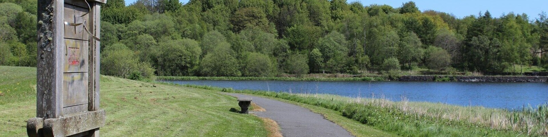 Un sendero pavimentado junto a un lago, con un cartel de madera y una mesa de picnic.