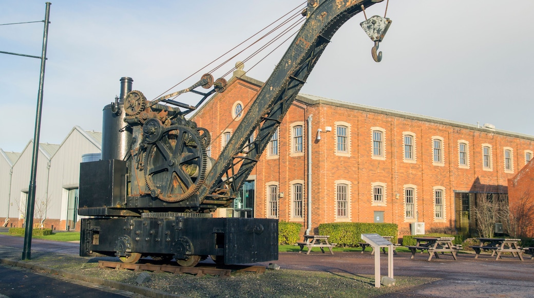 Une grande grue industrielle rouillée, dotée d’engrenages et d’un crochet, se dresse devant un bâtiment en briques comportant de nombreuses fenêtres.