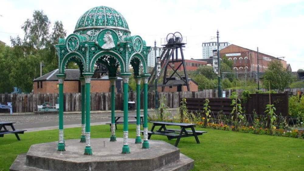A gazebo with a green dome and intricate designs, surrounded by a grassy area with picnic tables.