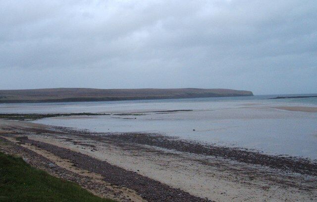 A coastal landscape with a sandy beach, calm water, and distant hills.