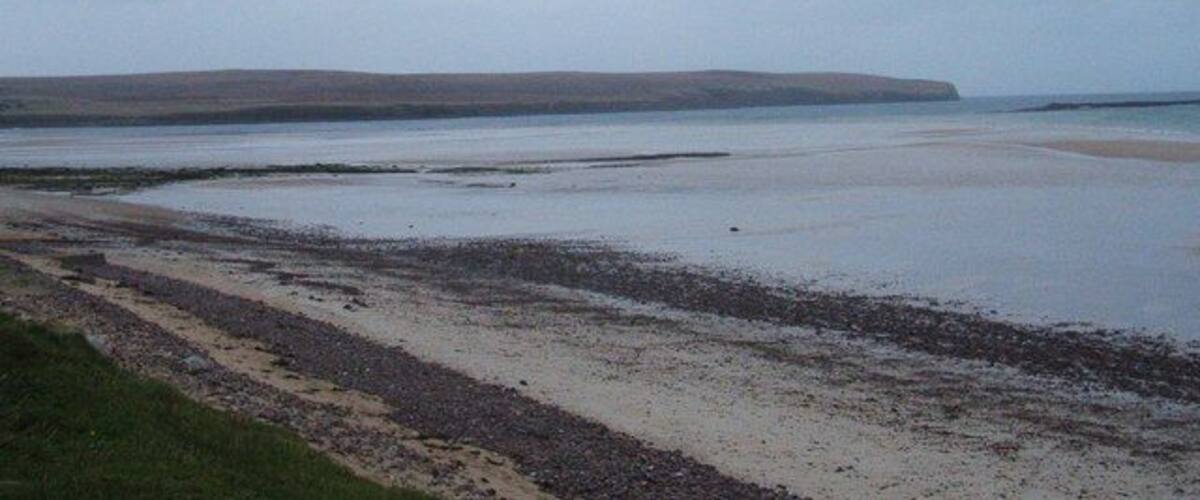 A coastal landscape with a sandy beach, calm water, and distant hills.