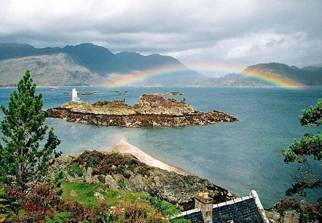 A coastal scene with a lighthouse, a rainbow, and mountains in the background.