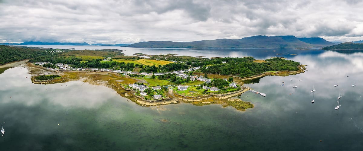A coastal village with houses and boats on calm waters, surrounded by mountains and greenery.