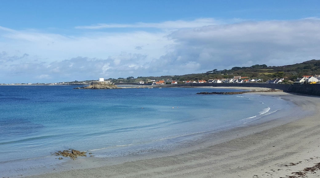 Uma praia costeira com águas azuis cristalinas, uma faixa de areia e uma pequena ilha com uma construção ao longe.