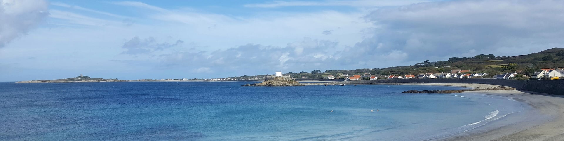 A coastal beach with clear blue waters, a sandy shore, and a small island with a structure in the distance.