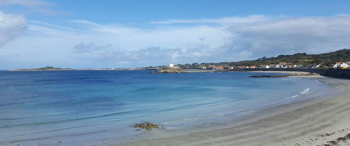 A coastal beach with clear blue waters, a sandy shore, and a small island with a structure in the distance.