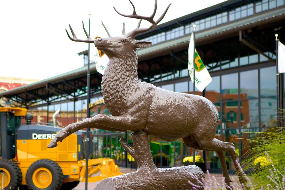 A deer statue in front of a building with flags and a yellow construction vehicle in the background.